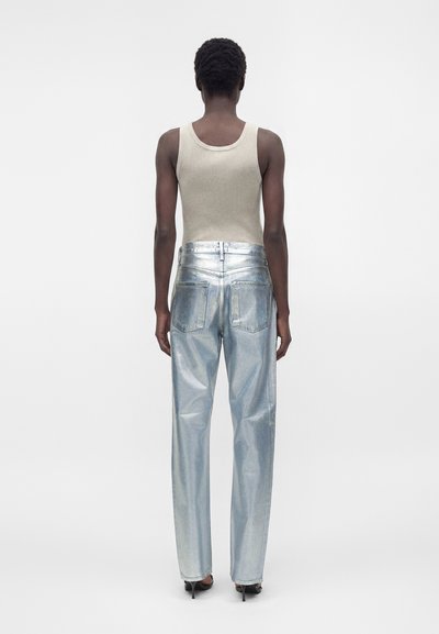 Άτομο που στέκεται με την πλάτη, φοράει μπεζ αμάνικο τοπ, μεταλλικό ασημί παντελόνι και μαύρα παπούτσια με ψηλό τακούνι σε απλό φόντο.