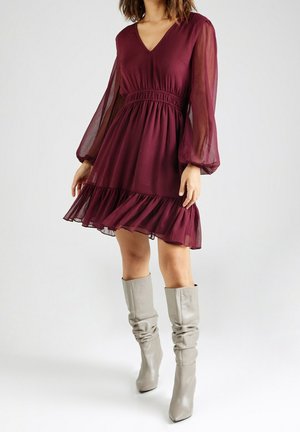 Robe bordeaux avec un décolleté en V et des manches longues en tissu transparent, présentant une taille froncée et un ourlet évasé. Associée à des bottes grises montant jusqu'aux genoux.