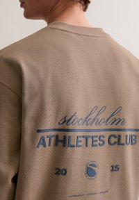 Brązowa bawełniana bluza z napisem "stockholm ATHLETES CLUB" w kolorze niebieskim, z okrągłym logo i rokiem "2015" nadrukowanym na plecach.
