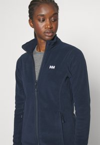 Marine fleecejack met hoge kraag, volledige ritssluiting aan de voorkant, twee zijzakken en geborduurd logo op de borst, met een gladde textuur.