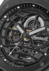 Zegarek z czarną czaszą w kształcie szkieletu o skomplikowanym wzorze, srebrnymi wskazówkami godzinową i minutową oraz złotymi akcentami. Widoczny wewnątrz mechanizm automatyczny.