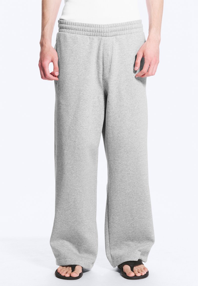 Homme portant un pantalon de survêtement ample gris clair avec taille élastique et tongs noires, debout les mains détendues le long du corps.