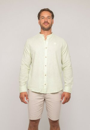 Homme debout, face à la caméra, portant une chemise à manches longues vert clair à boutons et un short beige, sur un fond uni.