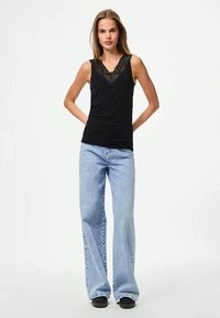 Top sem mangas preto com acabamento em renda na gola, combinado com jeans claros flare. Modelo a usar sapatos pretos, de pé com as mãos na anca.