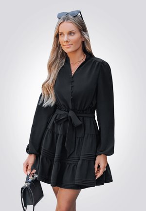 Vestido negro de manga larga, con botones en la parte delantera y un cinturón de lazo. Cuenta con volantes en capas en el dobladillo y una textura de tela suave.
