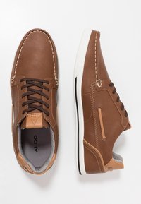 Chaussures décontractées en cuir marron avec semelles blanches, lacets noirs, intérieur gris rembourré, et logo ALDO à l'intérieur d'une chaussure.