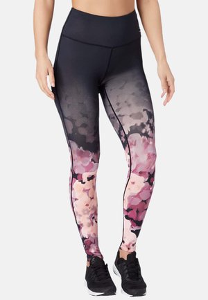 Woman wearing high-waist leggings with a black to pink floral gradient pattern and black athletic shoes on a white background.