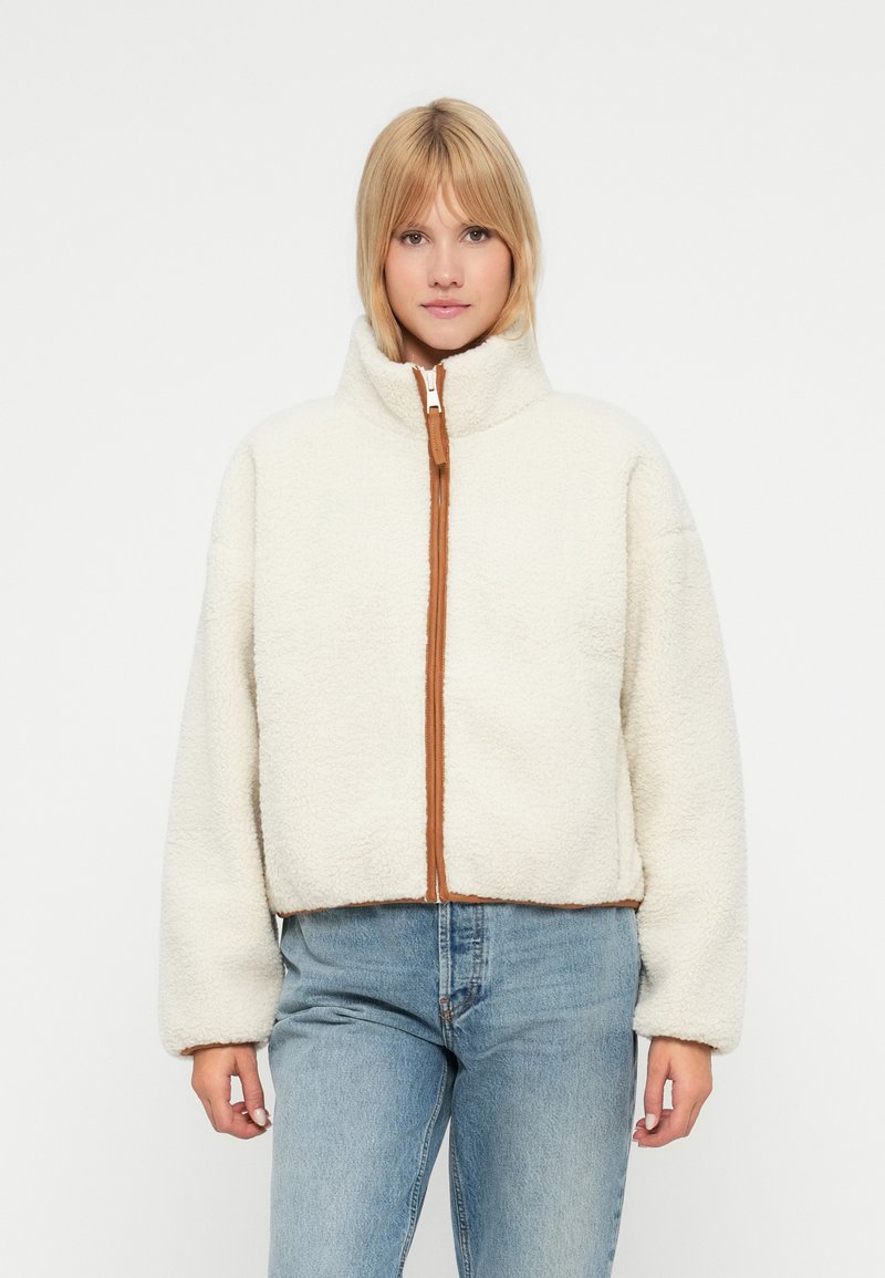 Bijela flis jakna s visokim ovratnikom, smešnim zatvaračem i skraćenim krojem. Mekana tekstura s rastezljivim manžetama i rubom.