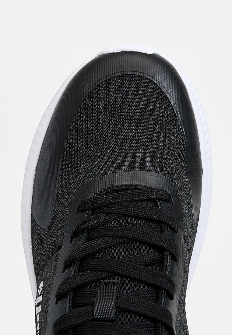 Zapatilla deportiva negra con parte superior de tela texturizada, suela blanca y cordones negros. Presenta una puntera suave y un branding sutil.