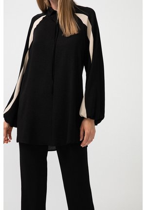 Vrouw die een zwarte tuniek draagt met een crèmekleurige streep op de mouwen en bijpassende zwarte broek, staand tegen een effen witte achtergrond.