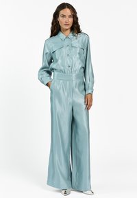 Vrouw met lang, krullend haar, gekleed in een glanzende lichtblauwe button-up shirt en bijpassende wijde broek, staande met één hand in de zak.