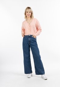 Cardigan rosa a maglia con scollo a V, caratterizzato da motivi a treccia e dettagli floreali, abbinato a jeans blu a vita alta con gamba larga e sneakers bianche.