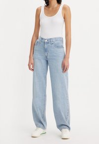 Femme portant un jean large bleu clair, un débardeur blanc, des baskets blanches et un collier argenté, debout devant un fond blanc uni.