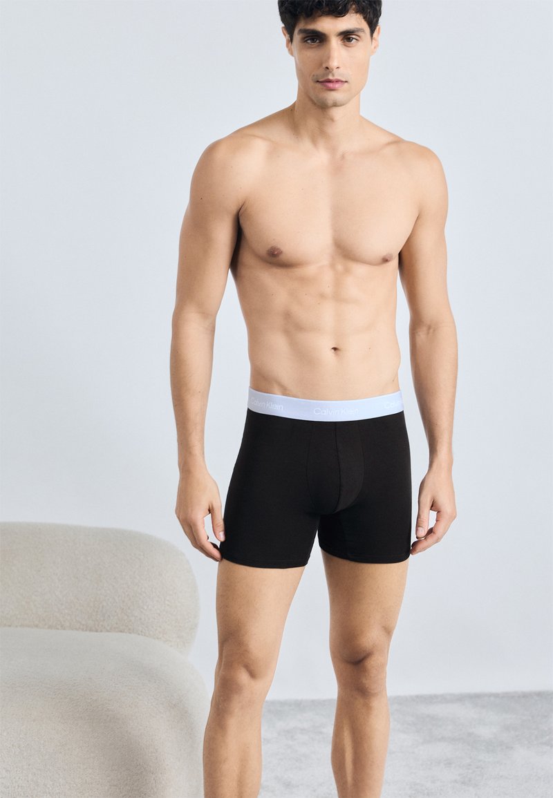 Čierne bavlnené boxerky s bielym elastickým pásom, na ktorom je logo "Calvin Klein". Dizajn je priliehavý a s dĺžkou po stred stehna.