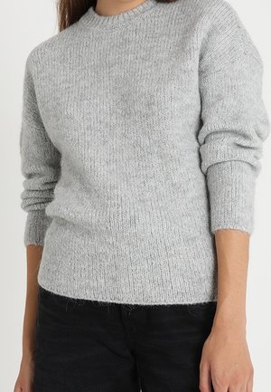 Frau trägt einen hellgrauen Strickpullover mit langen Ärmeln und schwarze Hose, steht vor einem schlichten Hintergrund.