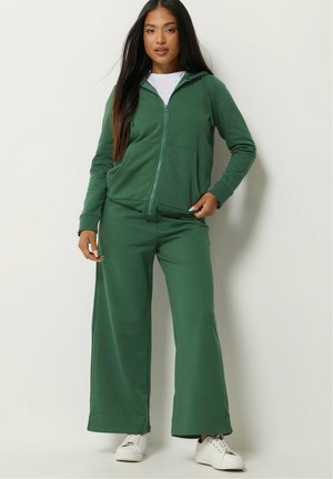 Femme portant un sweat à capuche vert zippé et un pantalon large avec des baskets blanches, se tenant devant un fond clair uni.