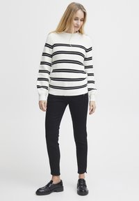 Sweater rayé en blanc et noir, texture tricotée, col rond ; associé à un pantalon slim noir et des mocassins noirs.