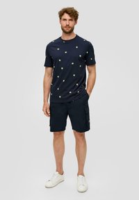 Mężczyzna stojący z rękami w kieszeniach, ubrany w granatową koszulkę z nadrukiem palm, ciemne szorty cargo i białe trampki na szarym tle.