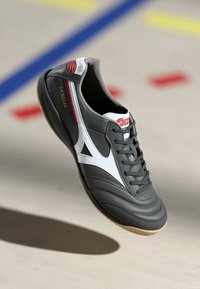 Pantofii de fotbal indoor gri cu accente albe, partea superioară din piele texturată, talpă din cauciuc gumă și un branding proeminent pe lateral și pe limbă.