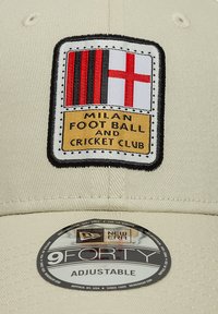 Beige baseball cap featuring a rectangular embroidered patch with red and black stripes, a white cross, and text reading "MILAN FOOT BALL AND CRICKET CLUB."