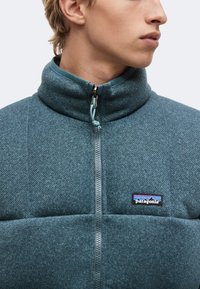 Νέος που φοράει τυρκουάζ fleece μπουφάν Patagonia με ψηλό φερμουάρ και κοιτάει δεξιά.