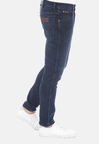 Jean skinny bleu foncé avec une texture en denim lisse, quincaillerie en ton cuivré et une patch en cuir marron sur la poche arrière.