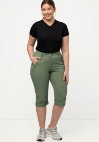 Pantaloni verdi fino al ginocchio con una texture liscia, tasche laterali e un design aderente, abbinati a una maglietta nera a maniche corte e sneaker grigie.