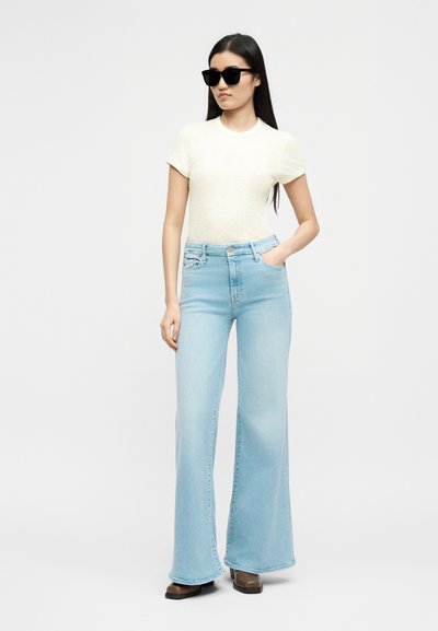 Γυναίκα που φοράει μαύρα γυαλιά ηλίου, κρεμ κοντομάνικη μπλούζα, ανοιχτό μπλε τζιν παντελόνι φαρδιές μπατζάκια και καφέ παπούτσια, στέκεται με το ένα χέρι στην τσέπη.