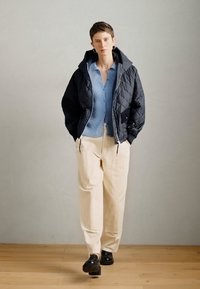 Gesteppte schwarze Jacke mit Kapuze, hellblauer gerippter Pullover, cremefarbene Hose und schwarze Schuhe. Glatte Textur, strukturierte Passform und kontrastierende Farben.
