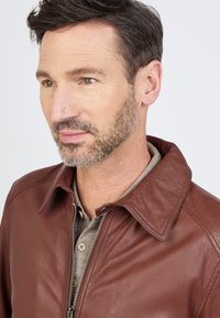 Braune Lederjacke mit einem klassischen Kragen und Reißverschluss, getragen über einem grauen Polo-Shirt mit Knopfdetails. Strukturiert.