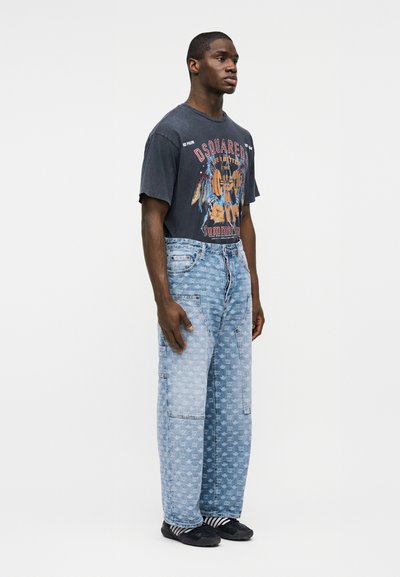 Άνδρας που στέκεται φορόντας μαύρο μπλουζάκι με σχέδιο, ανοιχτό μπλε φαρδιά τζιν με σχέδια, ριγέ κάλτσες και μαύρα παπούτσια slip-on.