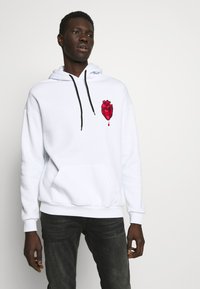 White hoodie with a red heart graphic. Features a front pouch pocket, black drawstrings, and ribbed cuffs and hem. Worn with dark jeans.
