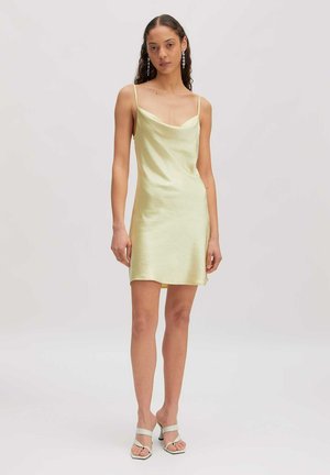 Frau steht in einem blassgelben Satin-Nachthemd mit dünnen Trägern, langen Ohrringen und weißen Sandalen mit Absatz vor einem einfachen hellen Hintergrund.