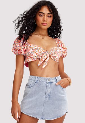 Vrouw met krullend haar, gekleed in een bloemenprint crop top met opgeblazen mouwen, voorgebonden aan de voorkant, en een lichtblauwe denim minirok, poserend tegen een effen achtergrond.