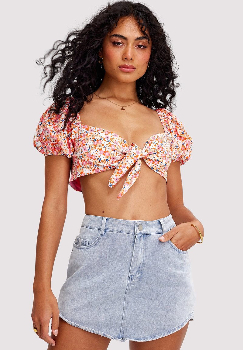 Femme aux cheveux bouclés portant un crop top à manches bouffantes à fleurs noué à l'avant et une mini-jupe en jean bleu clair, posant devant un fond uni.