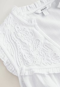 Chemisier blanc avec broderies texturées à motifs tourbillonnants, accents froncés sur les manches et finition en coton doux. Étiquette : NEXT.