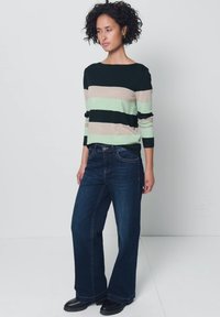 Vrouw met krullend haar, die een gestreepte trui en donkere wijde jeans draagt, staand tegen een effen lichte achtergrond.