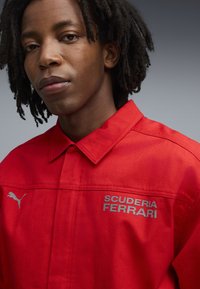 Cămașă roșie din bumbac cu guler cu nasturi. Prezintă un logo argintiu "SCUDERIA FERRARI" și logo-ul Puma pe piept. Textură netedă, croi casual.