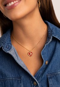 Collar de cadena de oro con un colgante en forma de corazón rojo y un centro texturizado, llevado sobre una camisa de mezclilla con diseño de cuello.