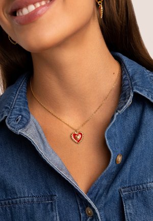 Collier en chaîne dorée avec un pendentif en forme de cœur rouge au centre texturé, porté sur une chemise en denim à col.