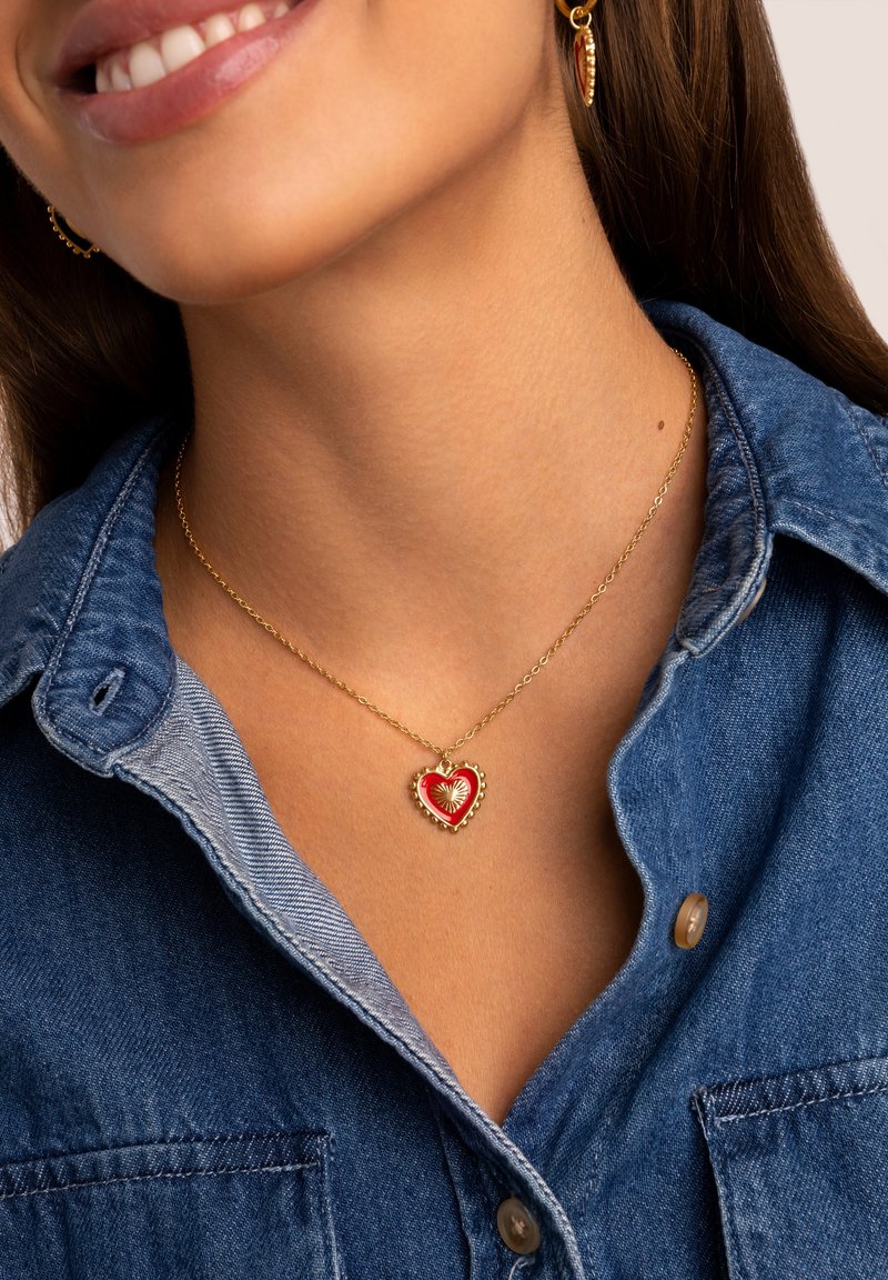 Collar de cadena de oro con un colgante en forma de corazón rojo y un centro texturizado, llevado sobre una camisa de mezclilla con diseño de cuello.