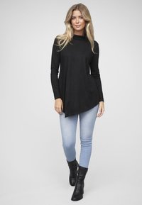 Zwarte top met lange mouwen en een colhals, met een asymmetrische zoom. De onderste randen zijn afgerond; gedragen met lichtblauwe skinny jeans en zwarte laarzen.