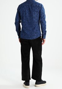 Sininen nappikauluspaita, jossa on lehtikuvio, yhdistetty mustiin corduroy-housuihin. Asuun kuuluu tummat kengät, jotka näkyvät takaa päin.