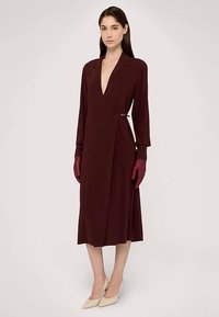Bordowa sukienka kopertowa z głębokim dekoltem w serek, długimi rękawami, bocznym ozdobnym klipsem i miękkim materiałem, zestawiona z czerwonymi rękawiczkami i beżowymi szpilkami.