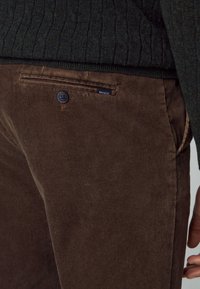 Pantalones de pana marrones con un bolsillo trasero abotonado y una pequeña etiqueta con el nombre de la marca. La tela tiene una apariencia texturada y estriada.