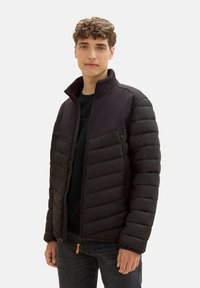 Młody mężczyzna w czarnej pikowanej kurtce z zamkami, stojący na jednolitym białym tle, patrzący w obiektyw.