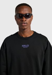 Mężczyzna w czarnych okularach przeciwsłonecznych i czarnej bluzie z fioletowym logo "HALO" na piersi, stojący na tle jednolitego szarego tła.