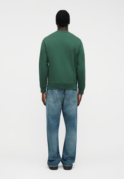 Άνδρας που φοράει σκούρο πράσινο φούτερ και φαρδιά μπλε τζιν παντελόνια, στέκεται με την πλάτη γυρισμένη στην κάμερα μπροστά σε ένα απλό λευκό φόντο.