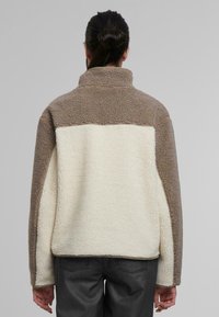 Chaqueta de forro polar en beige y crema. Cuello alto, mangas raglán, tejido texturizado. Presenta bloques de color contrastantes en la parte trasera.
