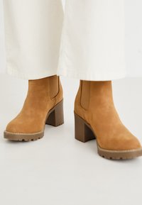 Botas de ante marrón hasta el tobillo con un tacón grueso, paneles laterales elásticos y una suela texturizada, combinadas con pantalones de pana ancha blancos.